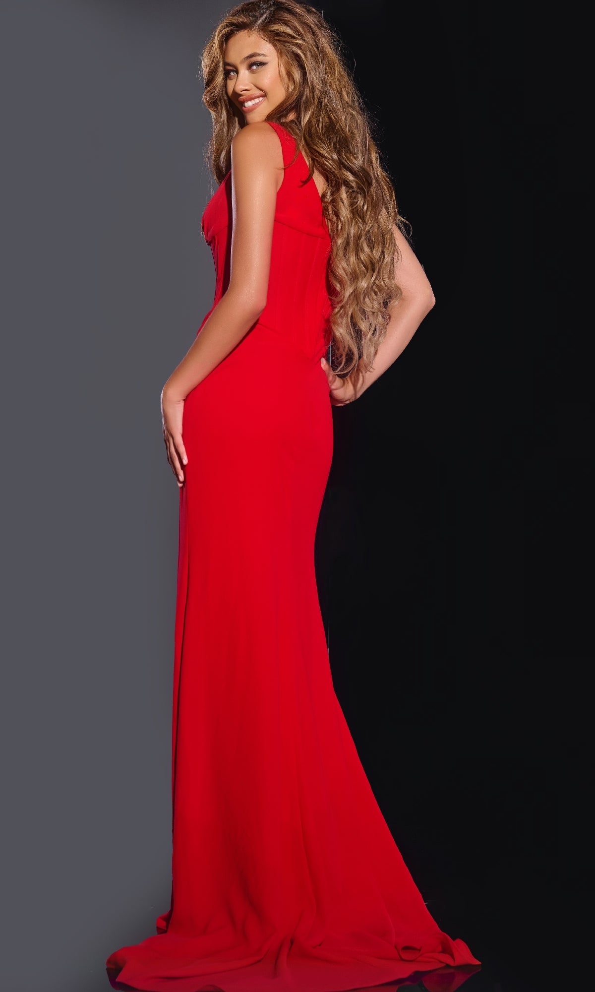 A woman with long wavy hair smiles over her shoulder, wearing the sleeveless Jovani 40783 long prom dress with a plunging v-neckline against a dark background.